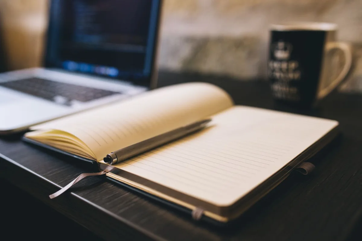 Open notebook with a pen on a wooden desk beside a laptop